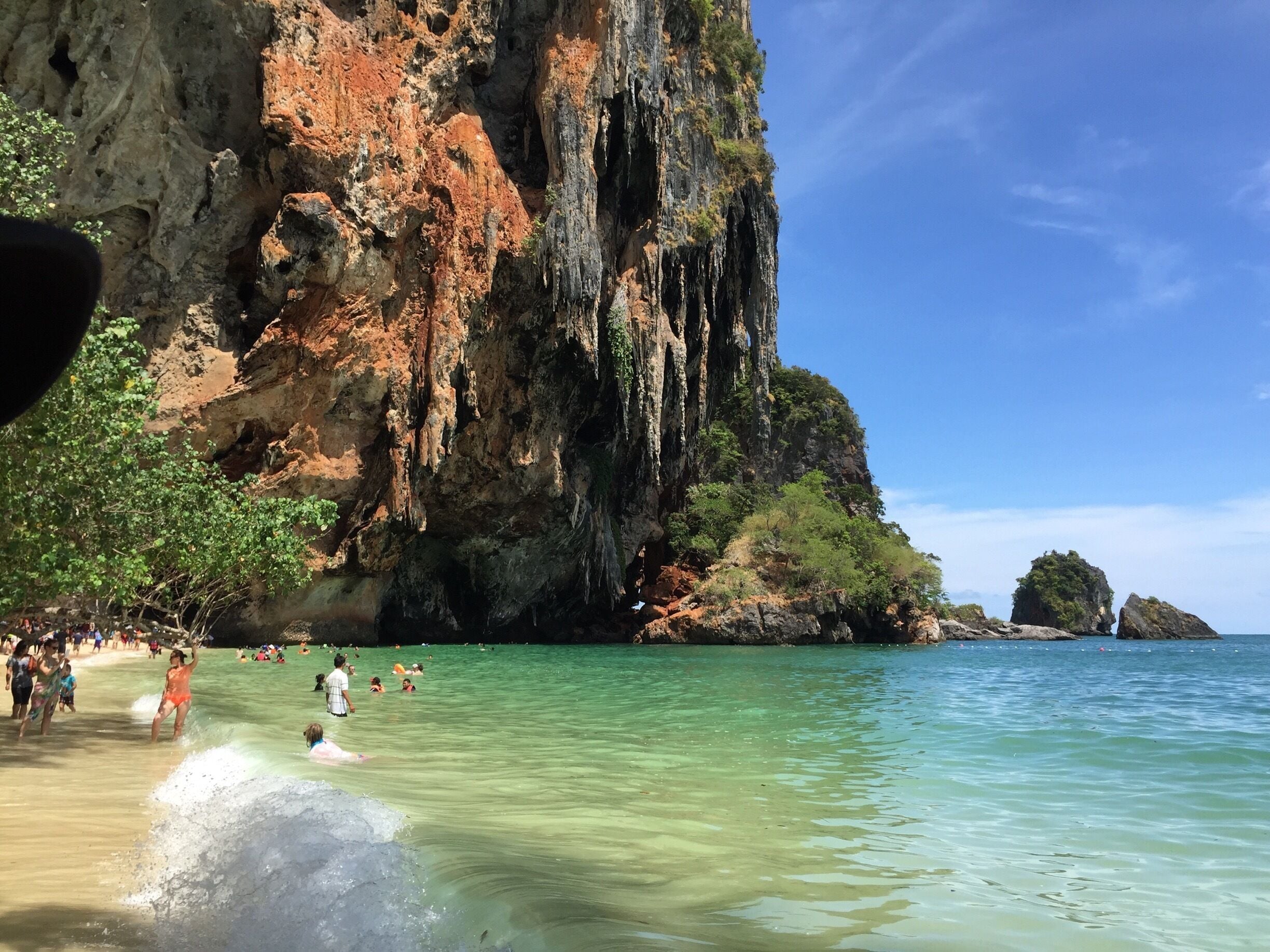 A coastal scene with clear turquoise waters, rocky cliffs, and a beach populated with people.