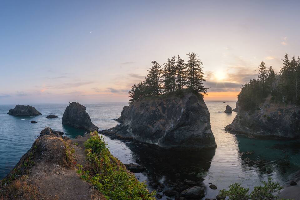 A coastal landscape with prominent rock formations, a sandy beach, and dense forested areas.