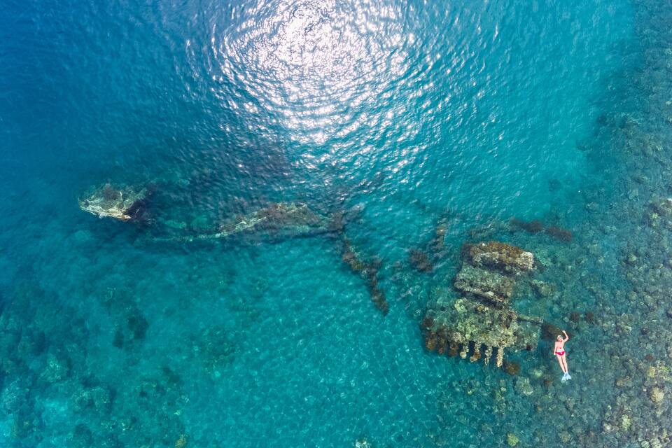 An aerial view of a clear blue sea with a large shipwreck and a diver on a rocky outcrop.