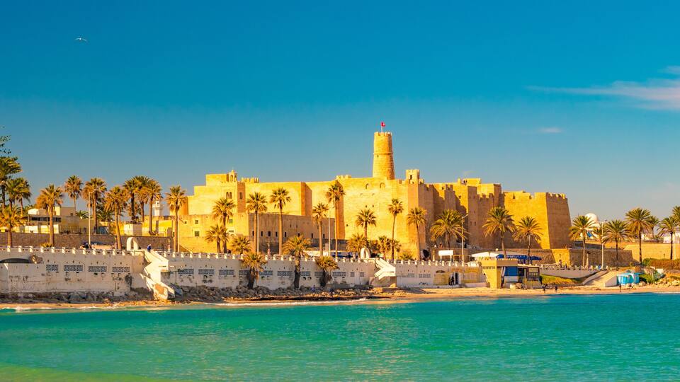 A coastal fortress with palm trees, a beach, and clear blue waters.