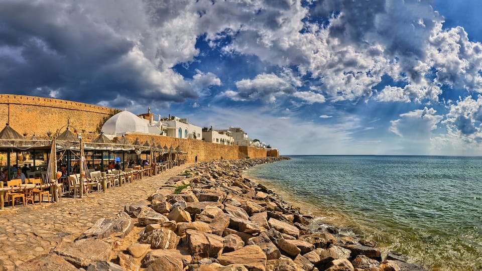 A coastal town with a stone wall, beachside café, and rocky shoreline.