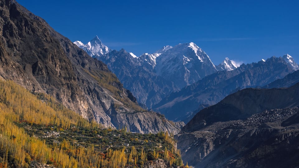 A mountainous landscape with snow-capped peaks, terraced fields, and a clear blue sky.