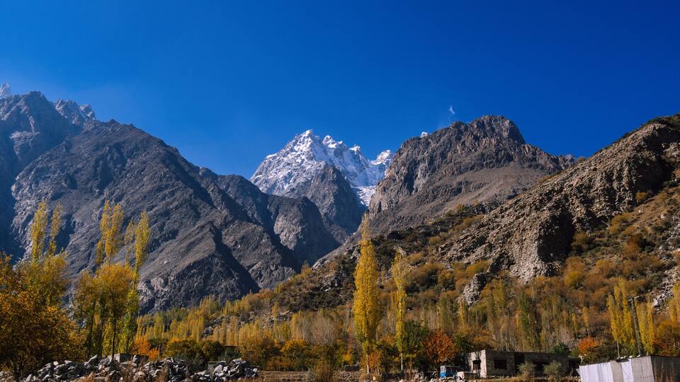 A mountainous landscape with snow-capped peaks, autumn-colored trees, and a small building in the foreground.