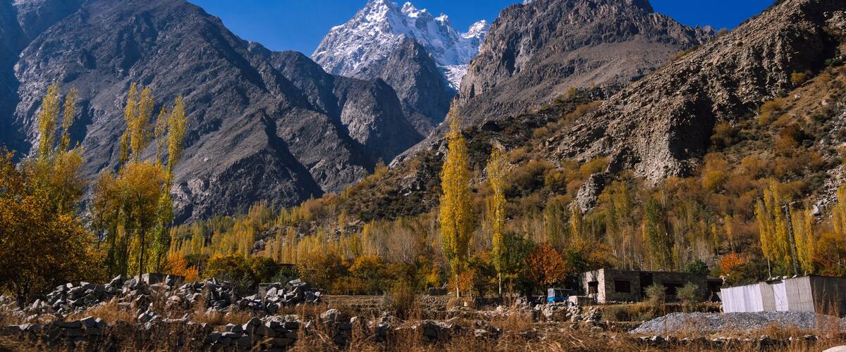 A mountainous landscape with snow-capped peaks, autumn-colored trees, and a small building in the foreground.