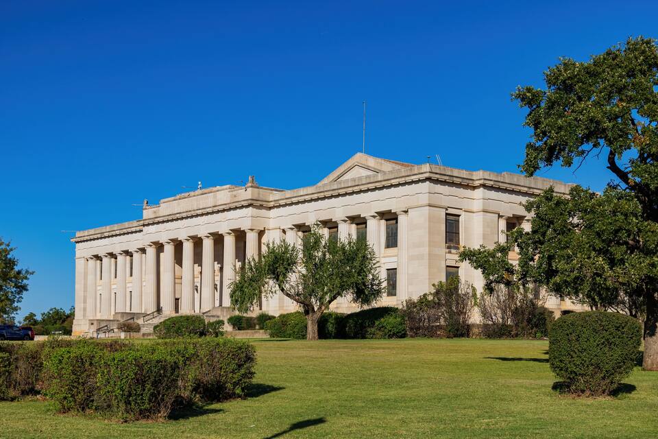 Un grand bâtiment de style néoclassique, doté de colonnes, entouré d’une pelouse bien entretenue et d’arbres.