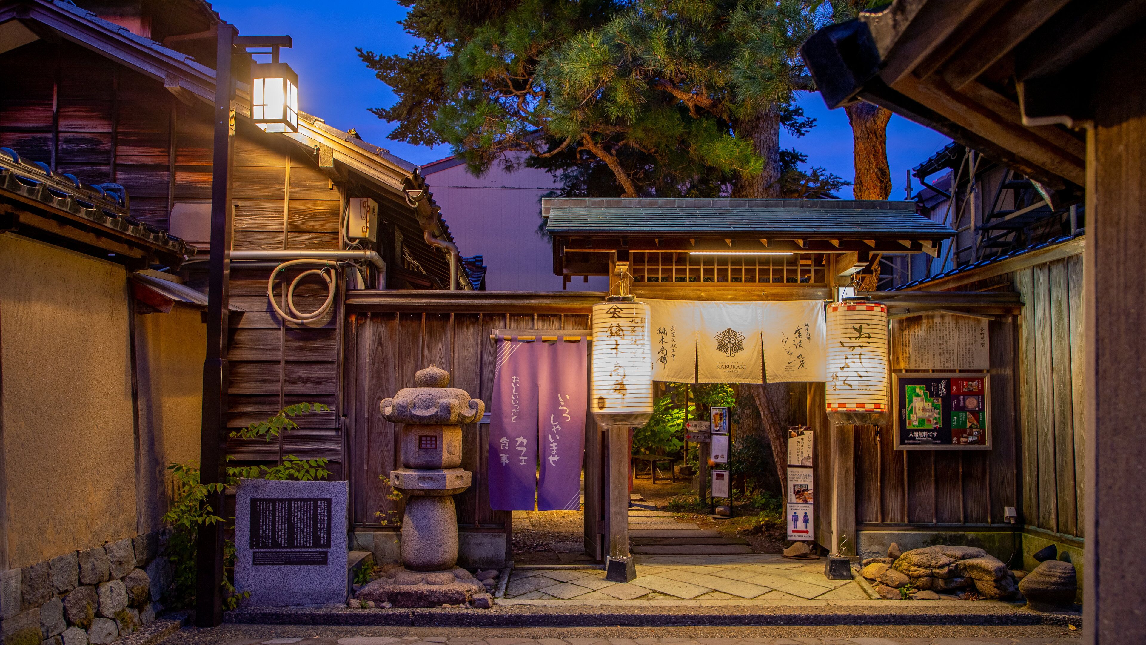 A traditional Japanese gate with lanterns and signage, surrounded by wooden structures and a stone lantern.