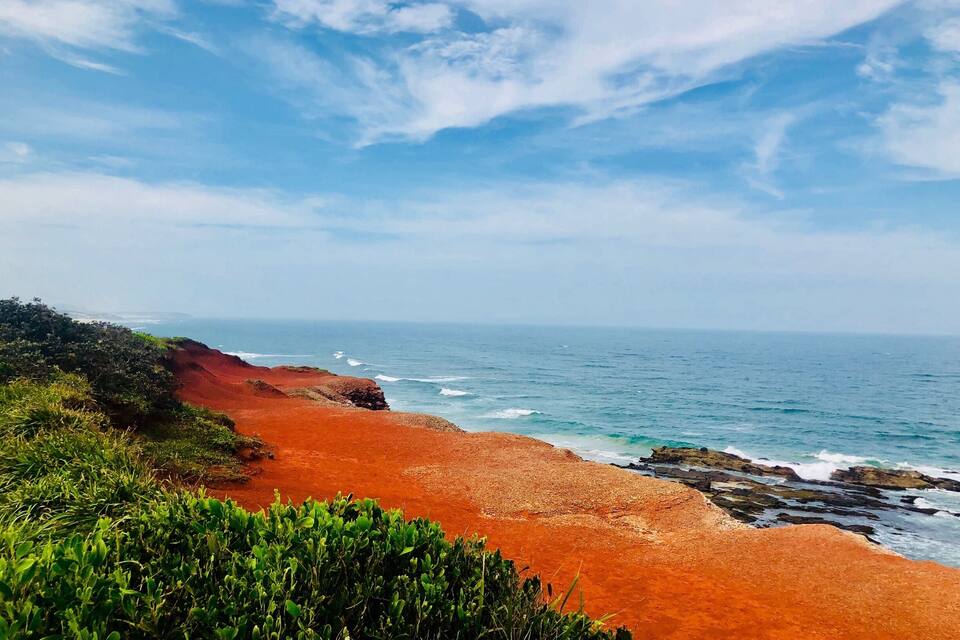 A coastal landscape with a red sandy beach, green vegetation, and the ocean under a partly cloudy sky.