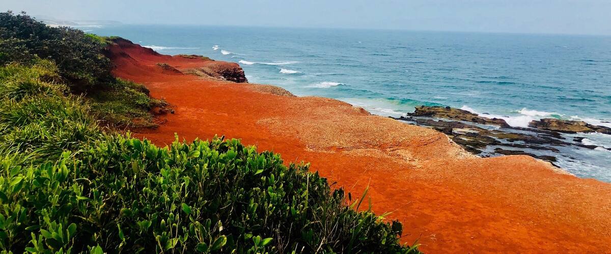 A coastal landscape with a red sandy beach, green vegetation, and the ocean under a partly cloudy sky.