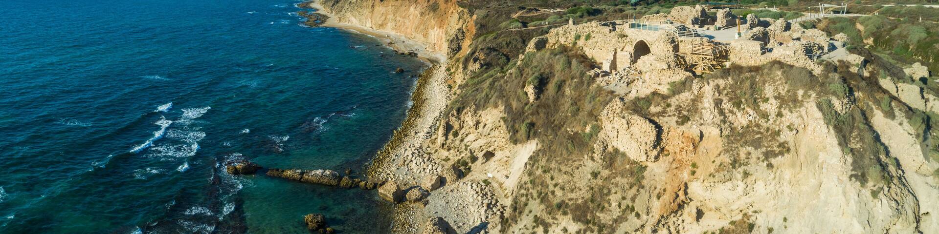 A coastal cliff with ruins, clear blue sea, and distant city skyline.