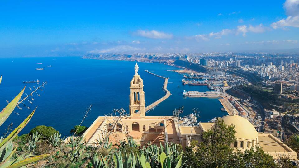 A coastal cityscape with a prominent church, a harbor, and a clear blue sea.