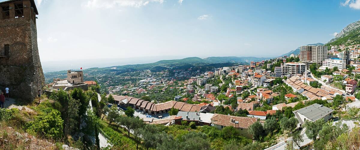 A historic stone tower, a hillside village with terracotta-roofed houses, and a mountainous backdrop.