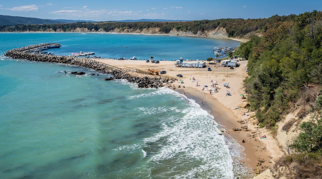A coastal beach with clear turquoise waters, a rocky breakwater, and a sandy shore with beachgoers.