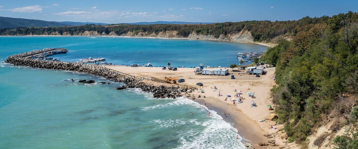 A coastal beach with clear turquoise waters, a rocky breakwater, and a sandy shore with beachgoers.