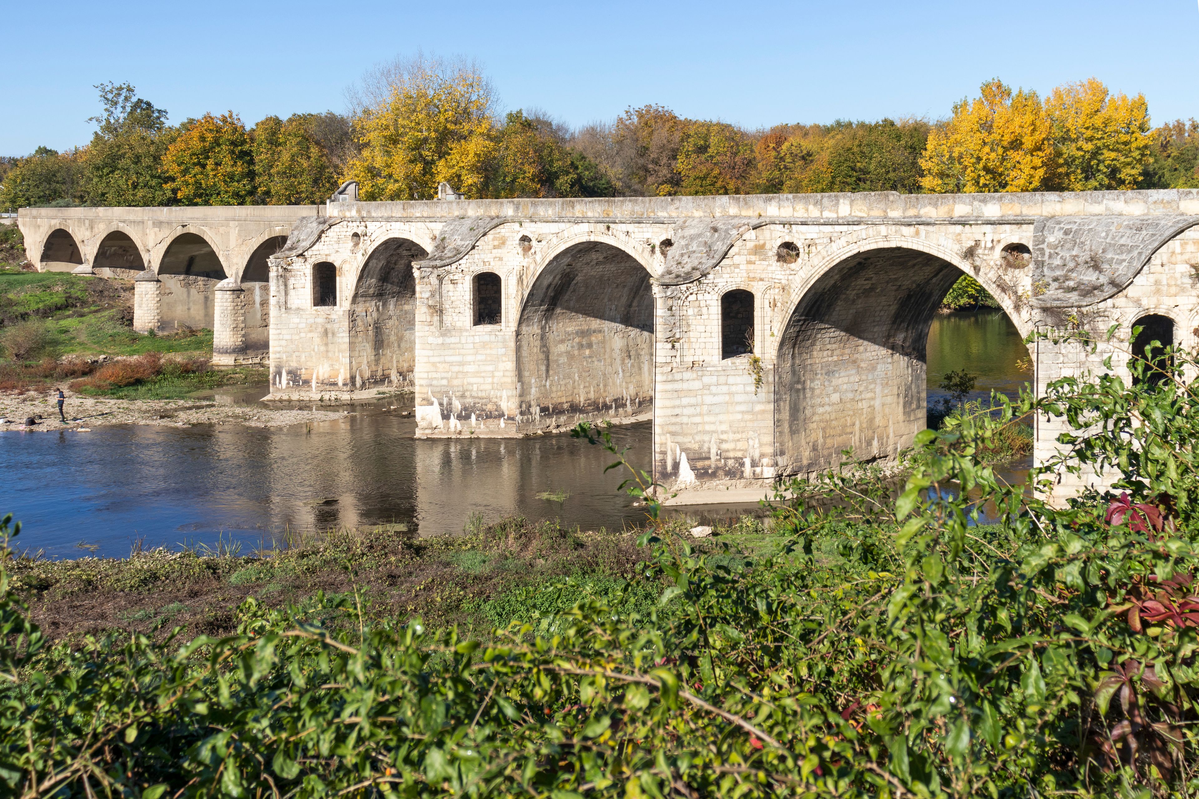 Un puente de piedra con múltiples arcos sobre un río, rodeado de follaje otoñal y vegetación.