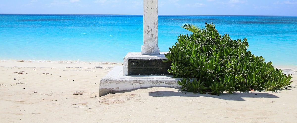 A white cross on a sandy beach with clear blue water and a blue sky.