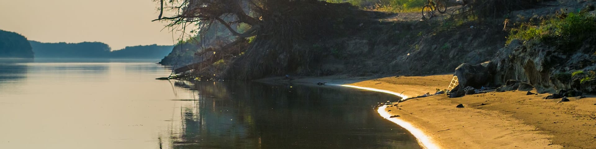 A calm lake with a sandy shore, surrounded by trees and a bicycle in the background.
