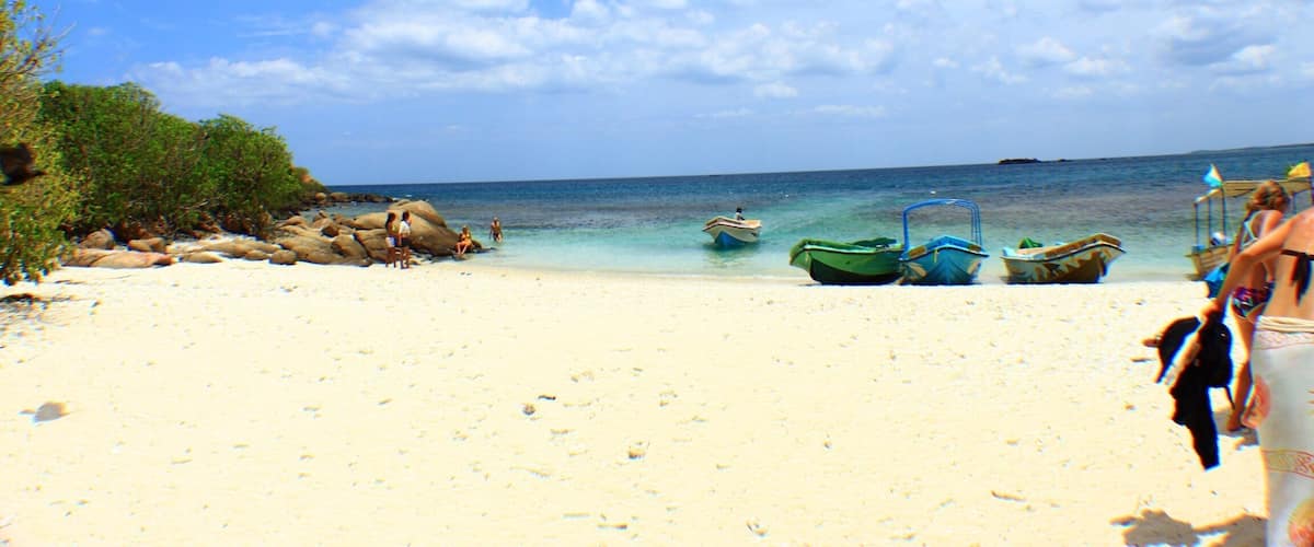 A beach with clear turquoise water, a few boats, and a rocky shoreline.