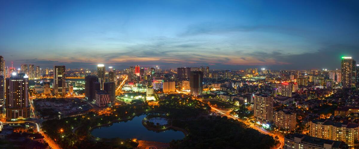 A cityscape at dusk with illuminated buildings and a central park with a lake.