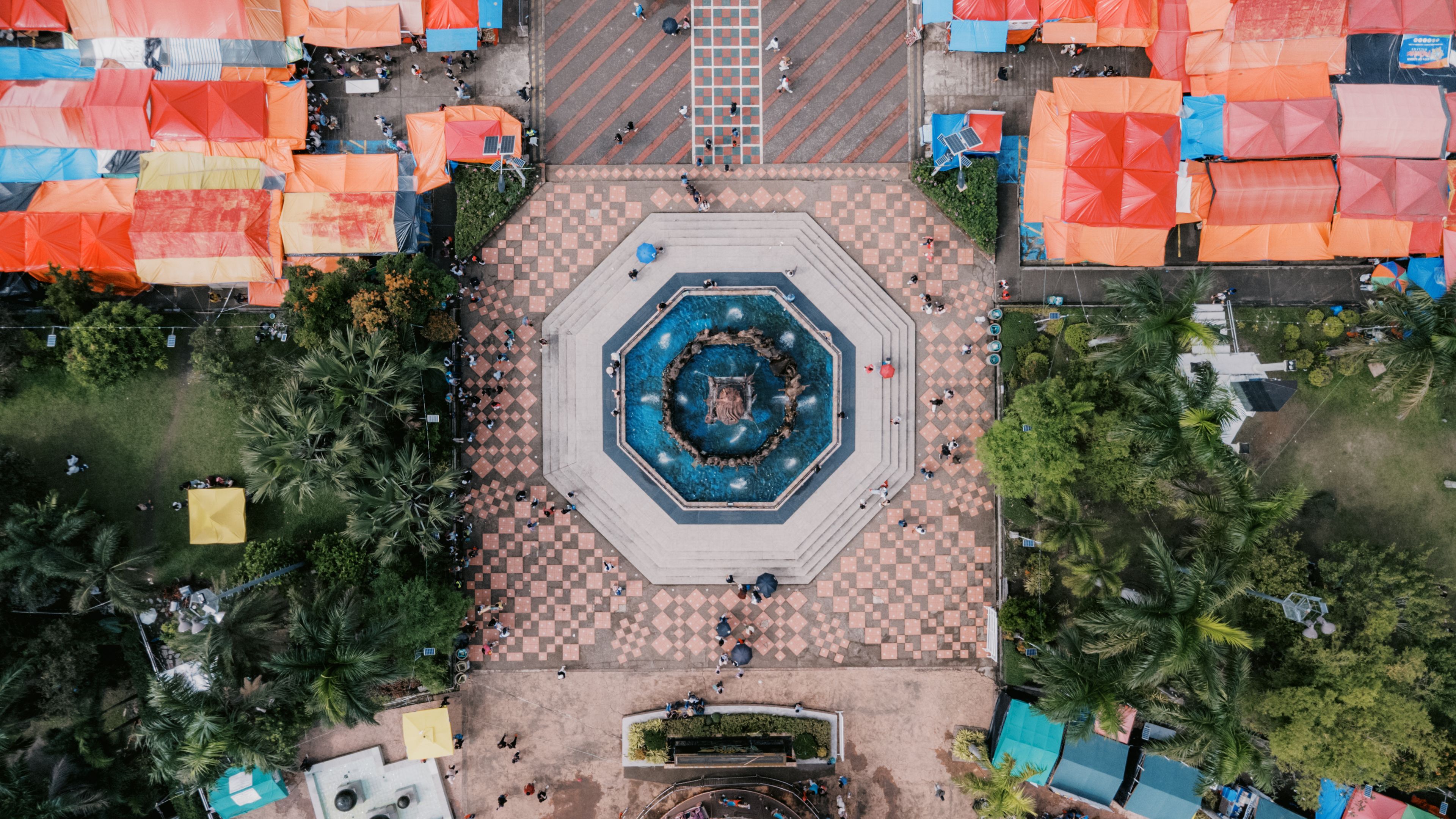 Vue aérienne d’une place avec une fontaine centrale, entourée de parasols orange et de palmiers.