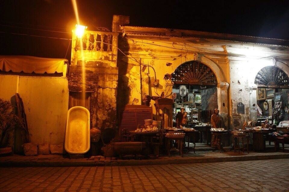 A nighttime market with a rustic building, a large archway, and various stalls.