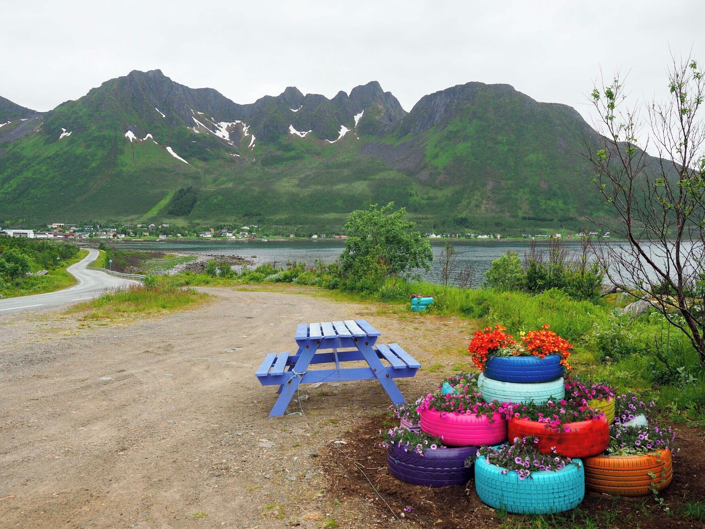 A scenic view of a lake with mountains in the background, a dirt path, a blue picnic table, and colorful tire planters with flowers.