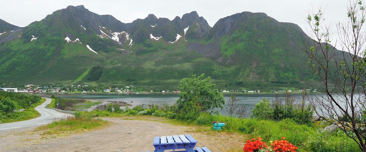 A scenic view of a lake with mountains in the background, a dirt path, a blue picnic table, and colorful tire planters with flowers.