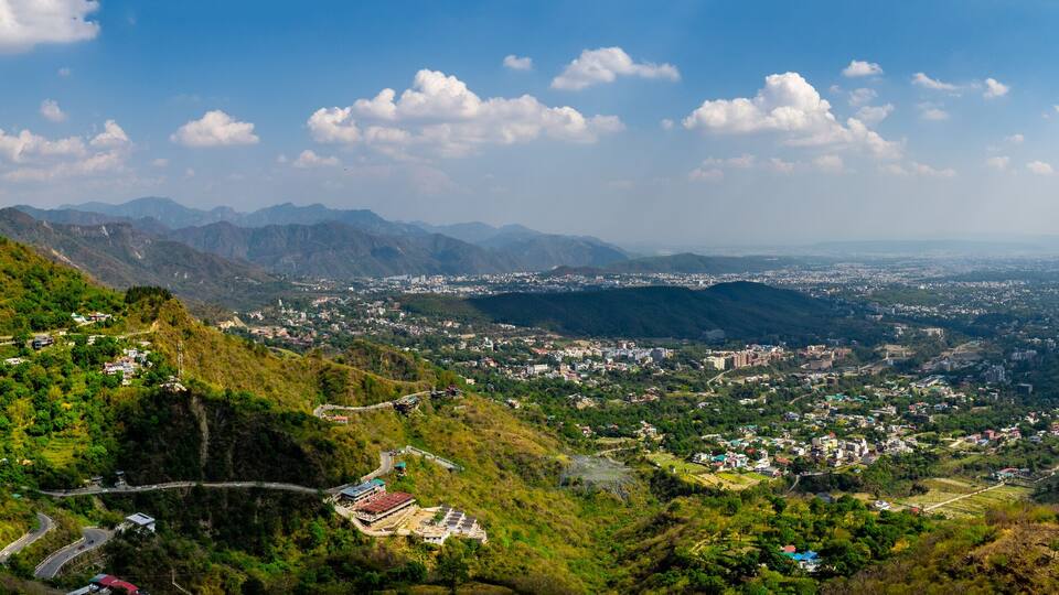 A mountainous landscape with a winding road, a valley with scattered buildings, and distant mountains under a partly cloudy sky.