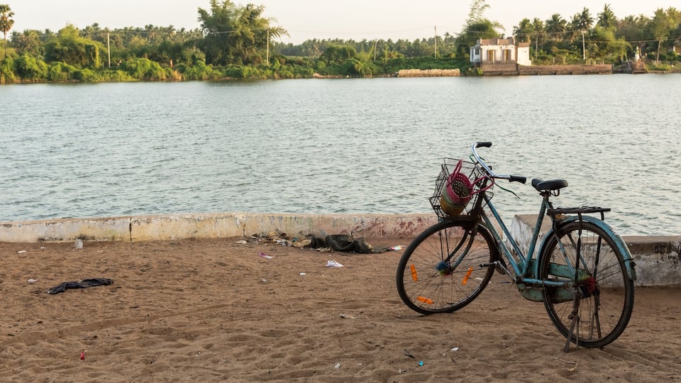 A bicycle parked on a sandy beach by a calm lake with greenery and buildings in the background.