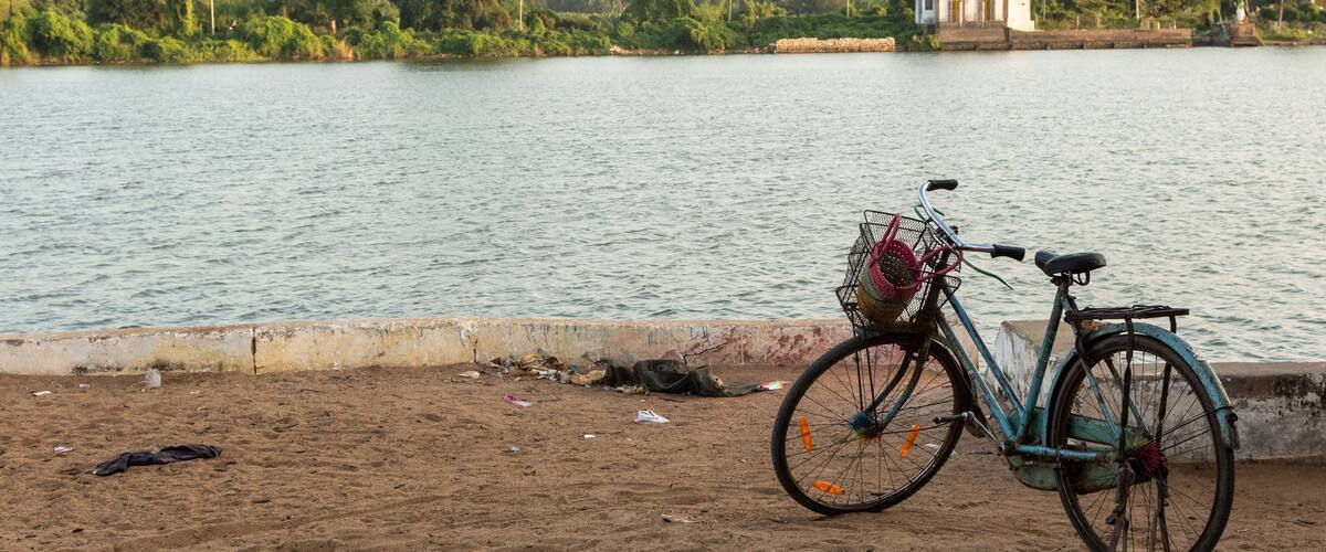 A bicycle parked on a sandy beach by a calm lake with greenery and buildings in the background.