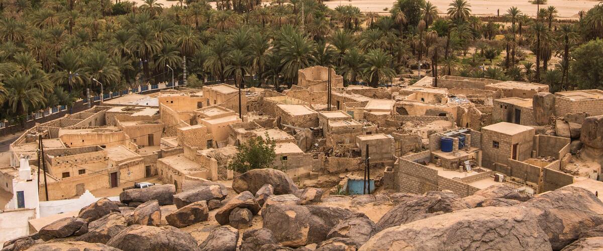 A desert village with traditional mud-brick buildings, palm trees, and a rocky mountain in the background.