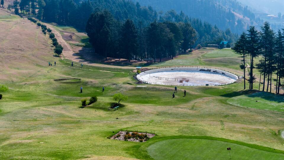 A golf course with a large circular green, a small pond, and a forested hillside.