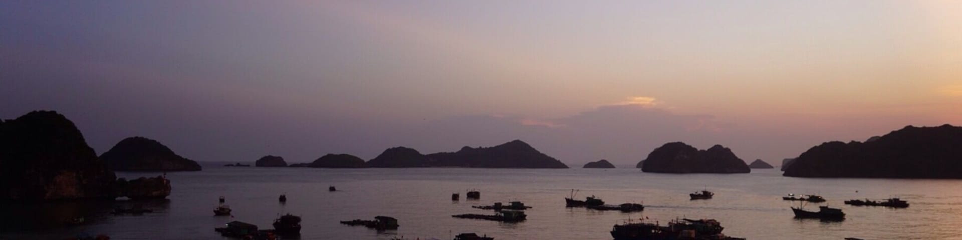 Cena costeira com vários barcos ancorados em águas calmas, rodeados por ilhas rochosas sob um céu crepuscular.