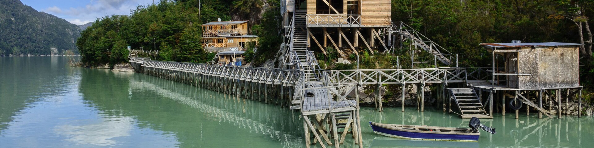 A wooden dock extending into a calm lake, surrounded by lush greenery and a mountain in the background.