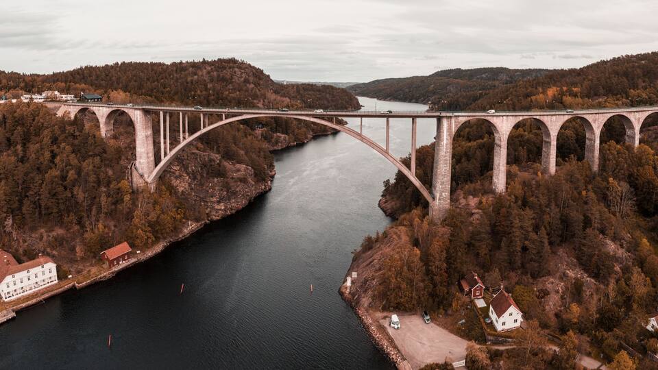 A large stone bridge spanning a river, surrounded by forested hills and a few buildings.