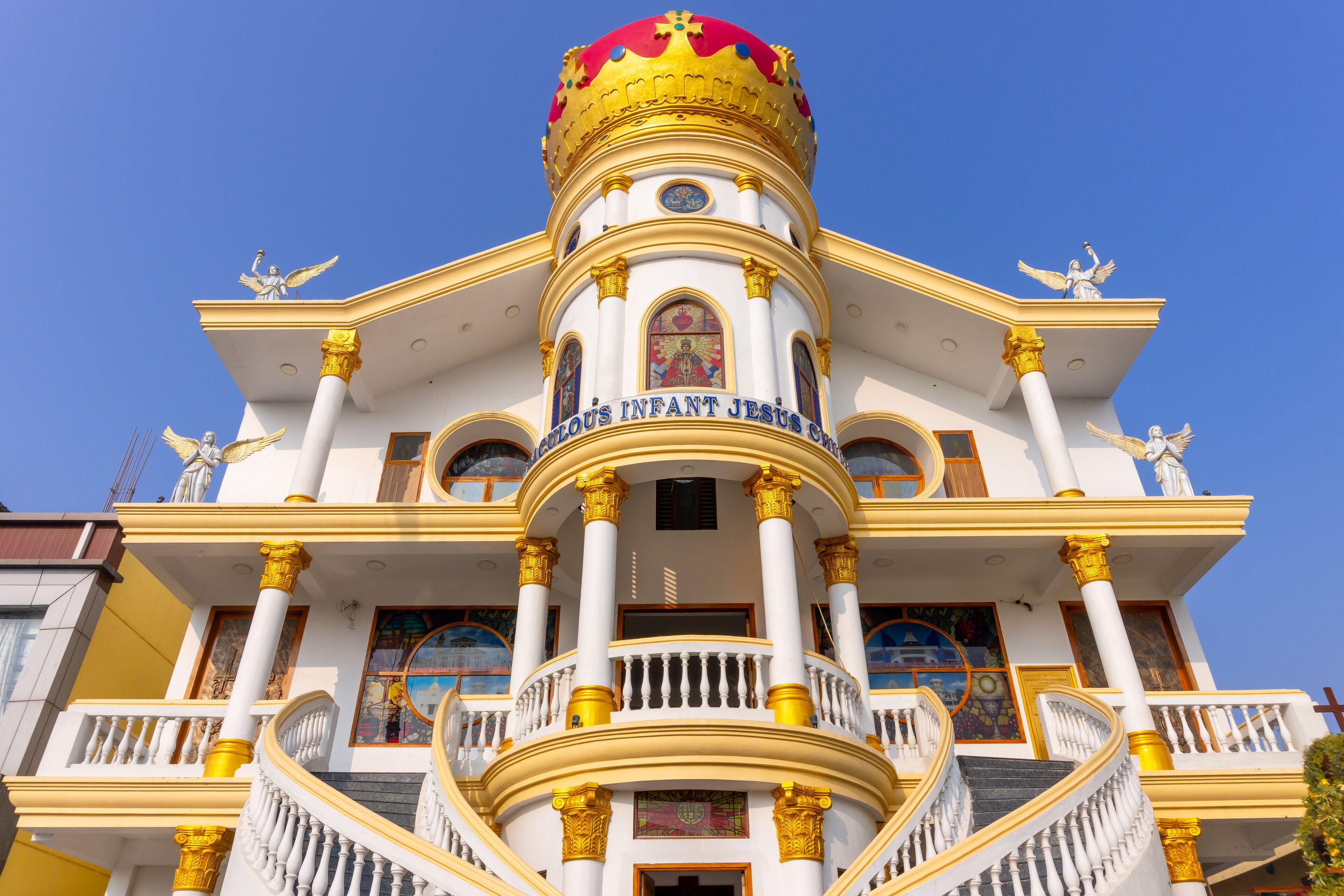 A white building with golden accents and a dome, featuring statues and the inscription "HOLY INFANT JESUS."