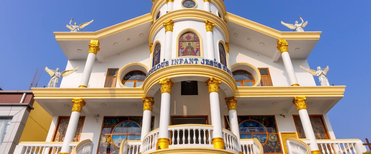 A white building with golden accents and a dome, featuring statues and the inscription "HOLY INFANT JESUS."