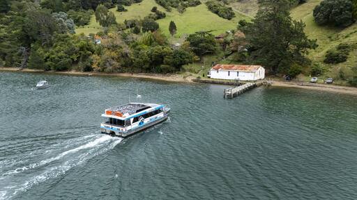 Aerial view of the Pelorus Mail Boat at Wilson Bay slicing through the dark water, leaving a frothy white wake against the backdrop of verdant hills, Marlborough Sounds, New Zealand.