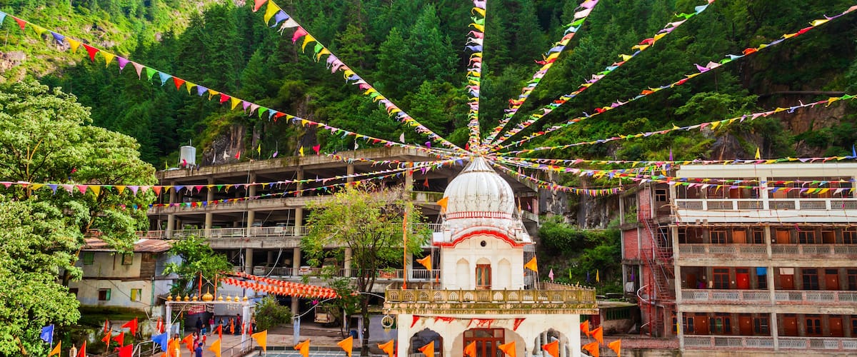 A temple with colorful prayer flags, a water body, and a forested mountain backdrop.