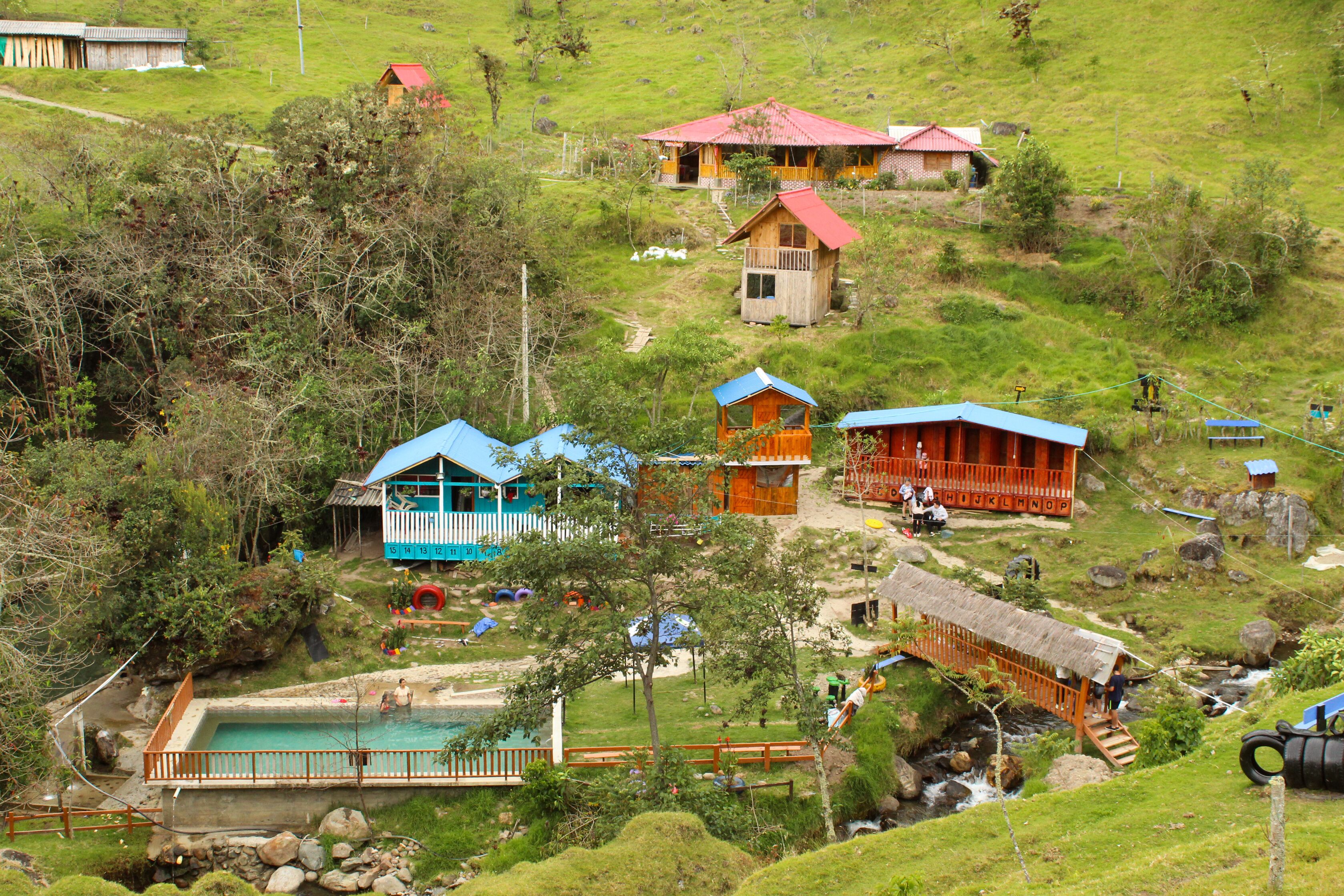 A hillside village with colorful wooden houses, a swimming pool, and a thatched bridge.