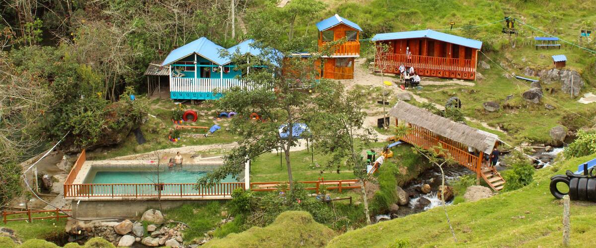 A hillside village with colorful wooden houses, a swimming pool, and a thatched bridge.