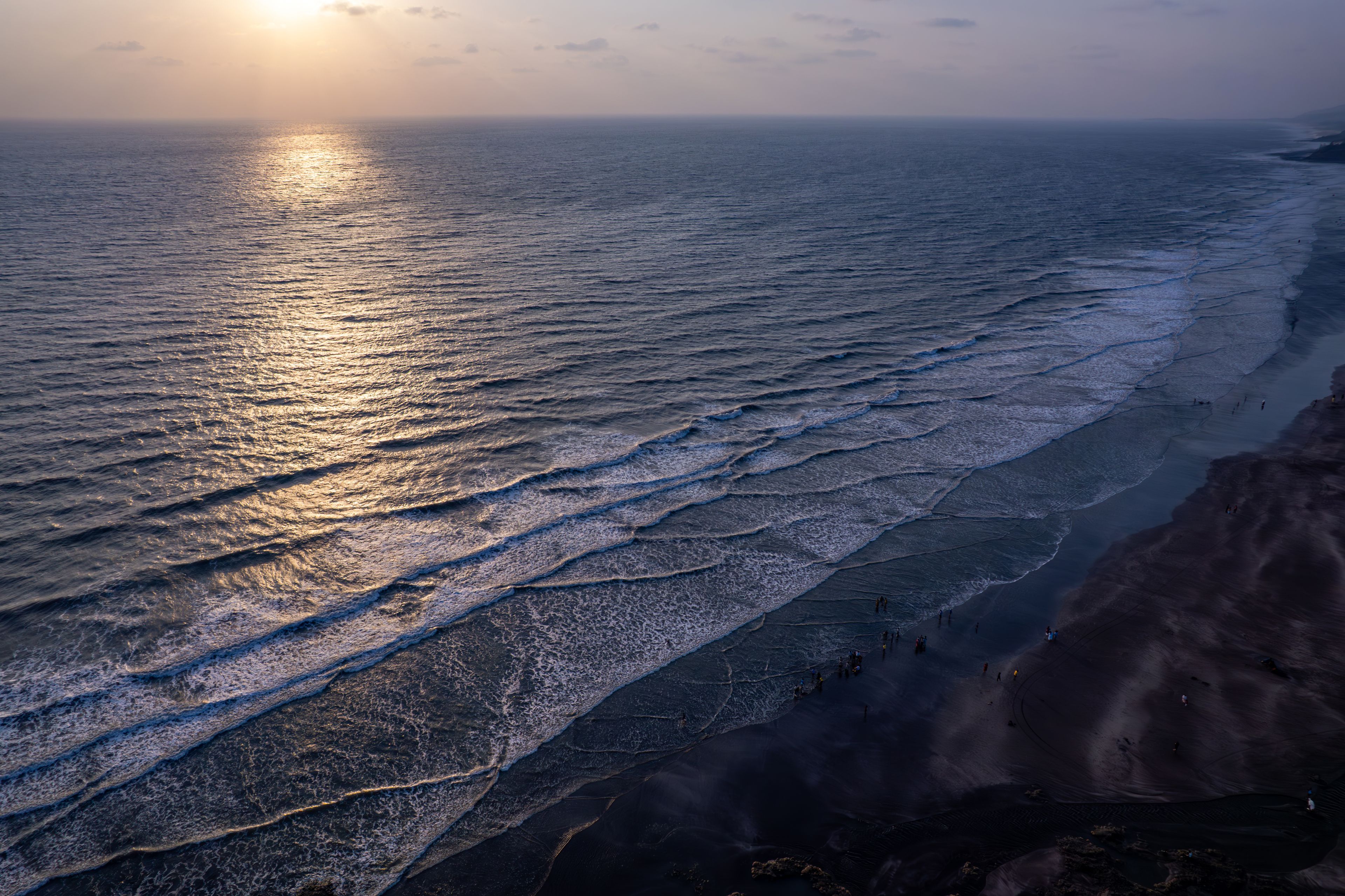 Paisagem costeira com ondas se aproximando da praia, céu claro e o sol se pondo ou nascendo perto do horizonte.