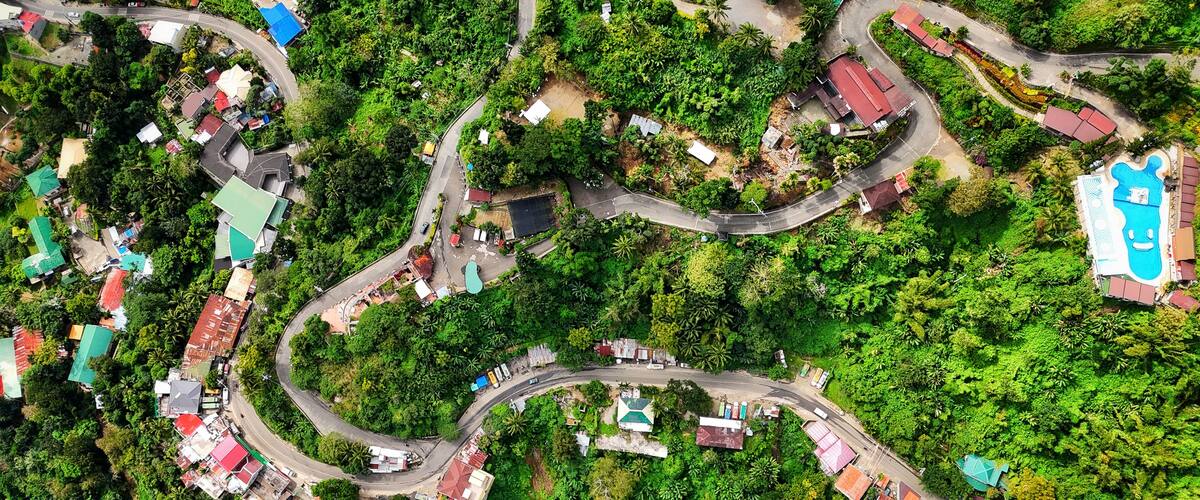 An aerial view of a winding road through a lush, green landscape with scattered buildings and houses.