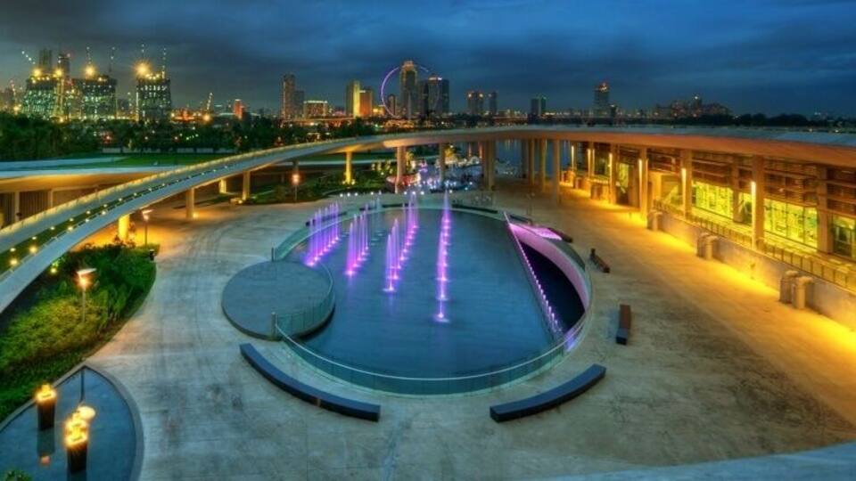 A waterfront promenade with a circular water feature and illuminated fountains.