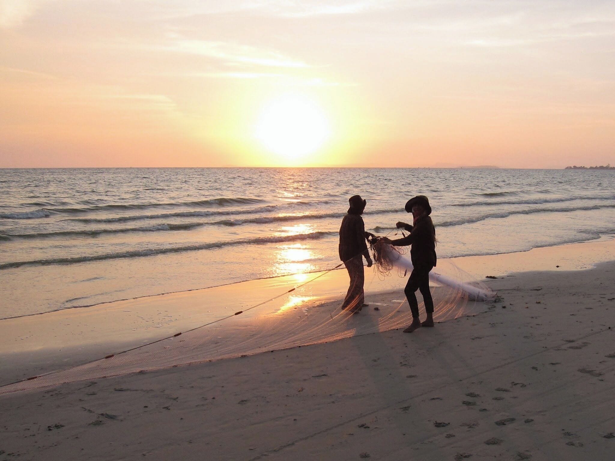 Two people fishing on a beach at sunset.