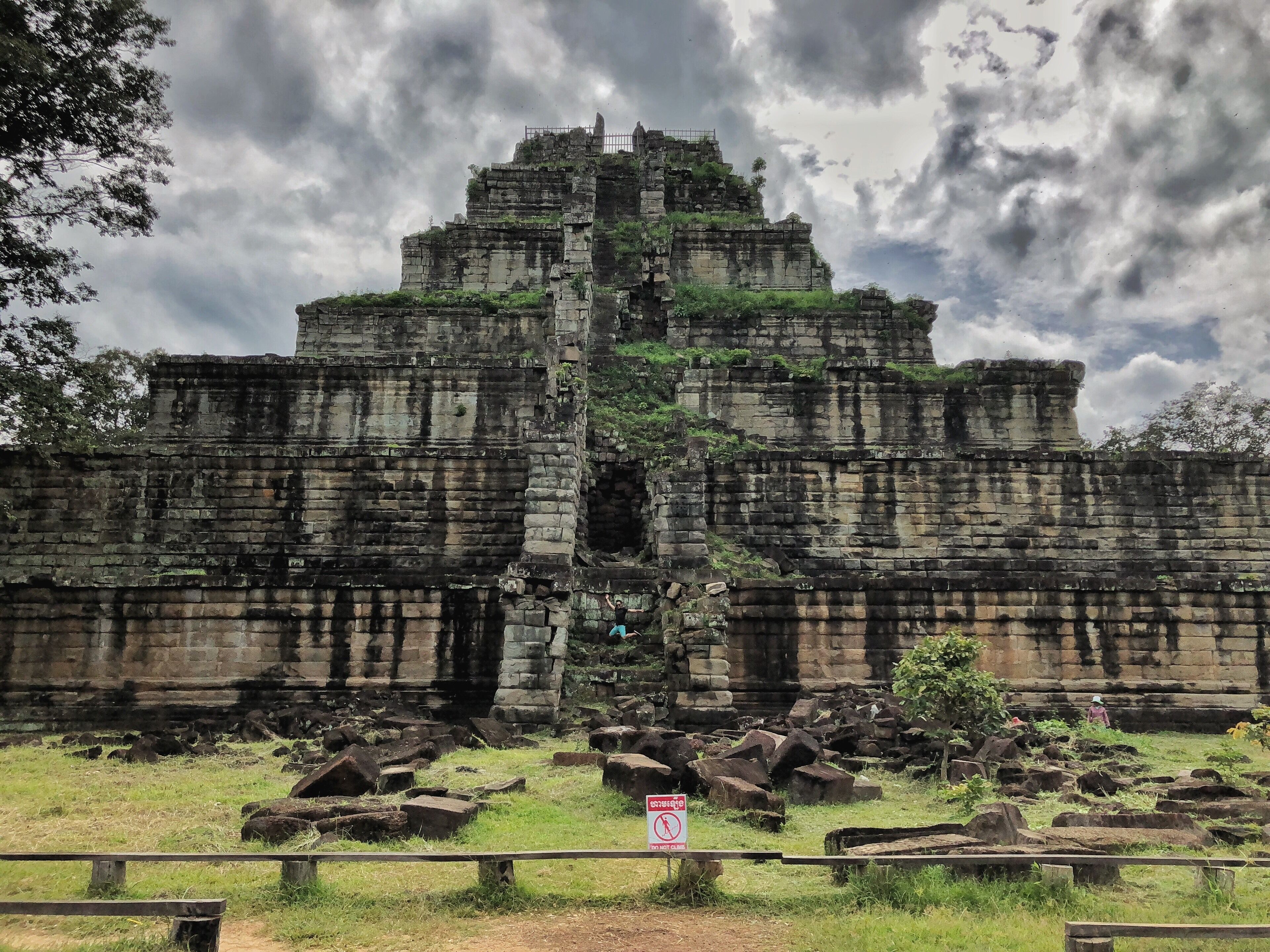 An ancient stone structure with a central tower and multiple levels, surrounded by scattered rocks and greenery.