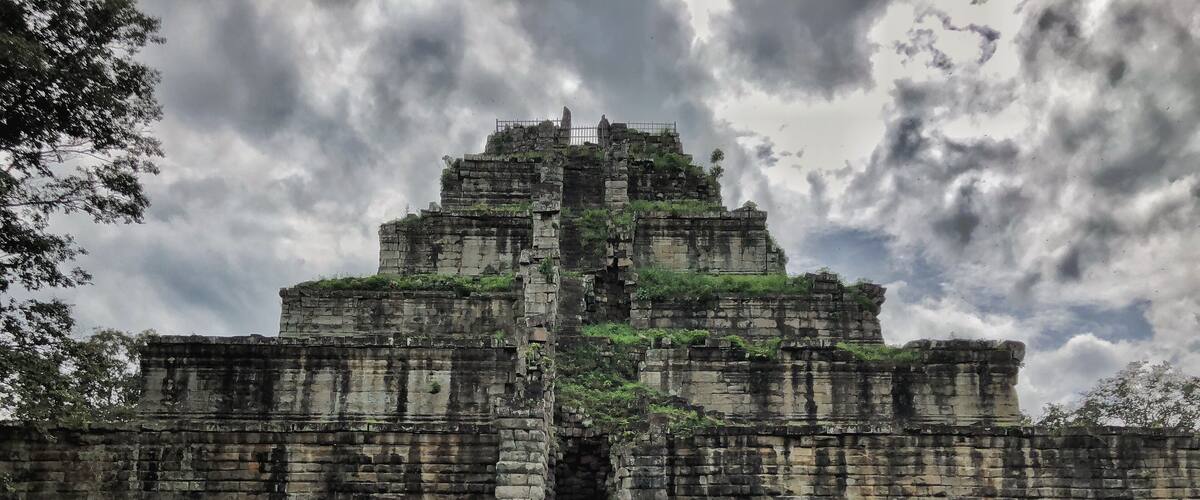 An ancient stone structure with a central tower and multiple levels, surrounded by scattered rocks and greenery.