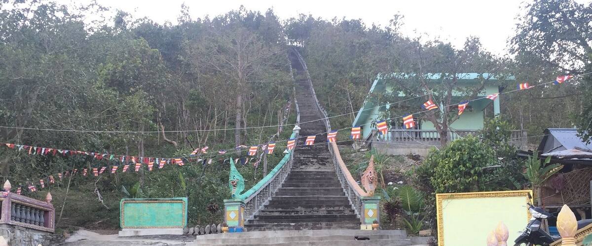 A staircase leading up to a temple with a green roof, surrounded by trees and decorated with flags.