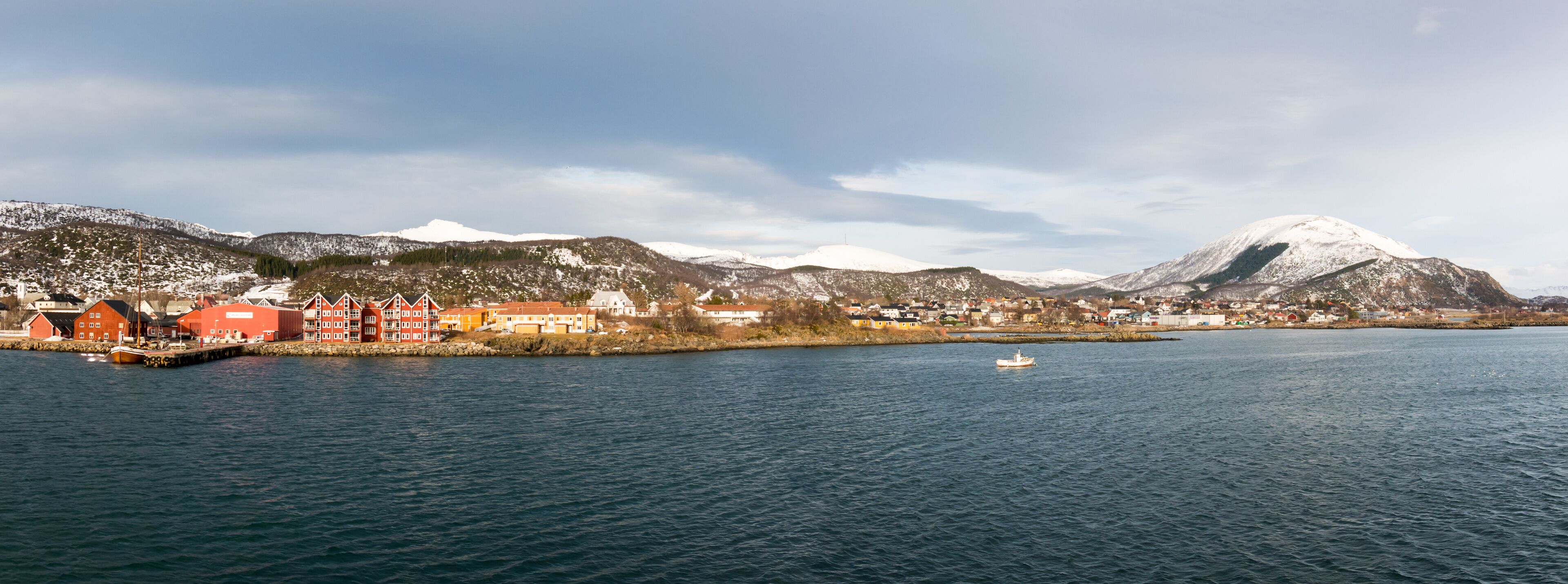 A coastal town with red buildings, a body of water, and snow-capped mountains in the background.