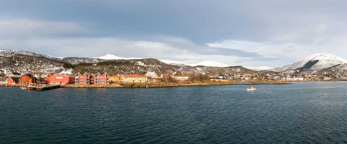 A coastal town with red buildings, a body of water, and snow-capped mountains in the background.