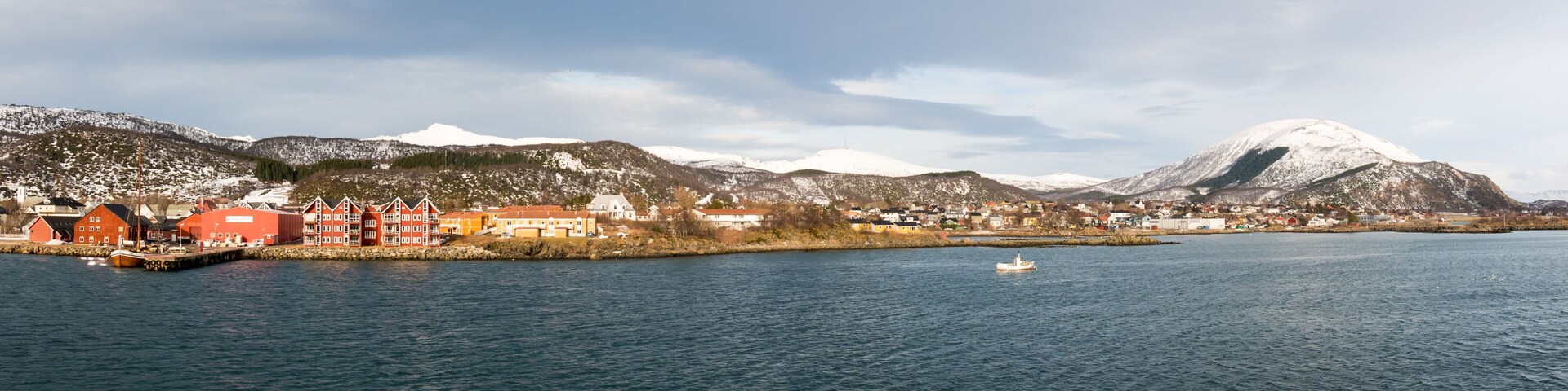 A coastal town with red buildings, a body of water, and snow-capped mountains in the background.